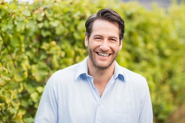 A smiling man in a light blue button-down shirt standing in a vineyard.