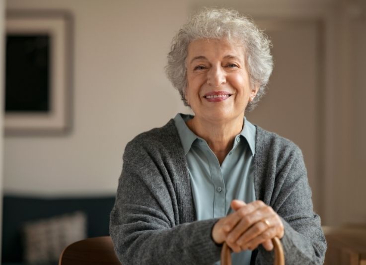 A smiling older woman with curly grey hair wearing a grey cardigan over a light shirt, resting her hands on the handle of a cane.